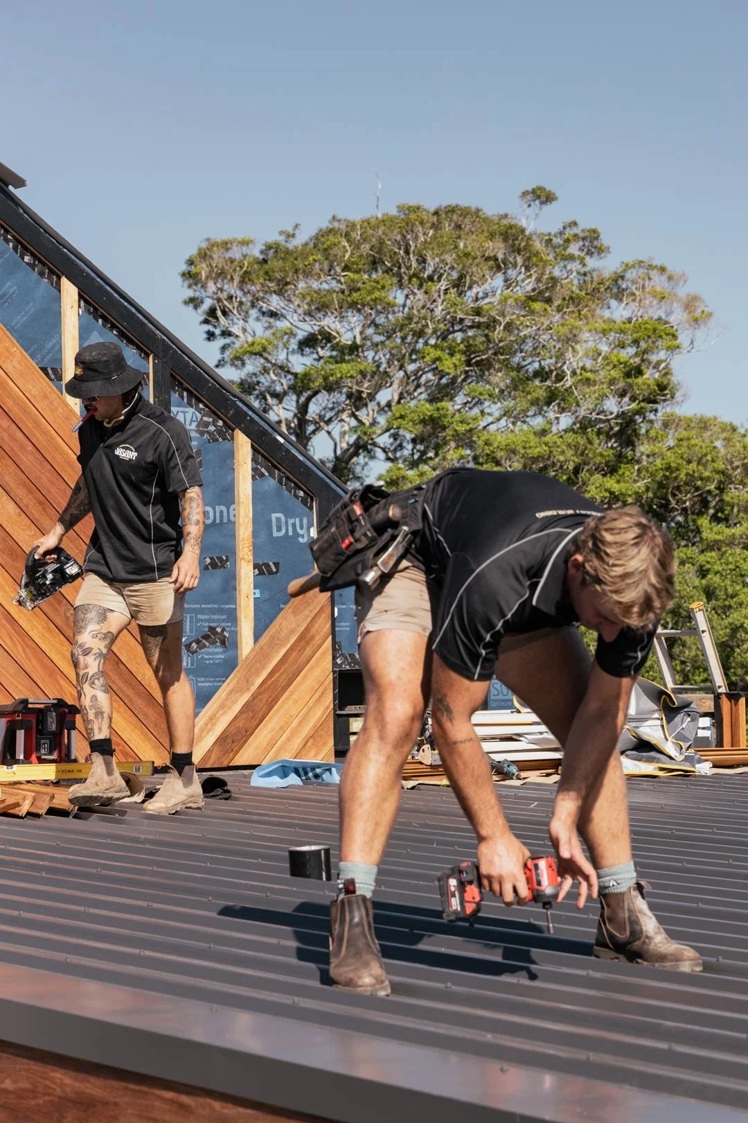 Two workers installing a metal roof on a building, with one bending down using a cordless drill and the other walking nearby, against a background of trees and clear sky.
