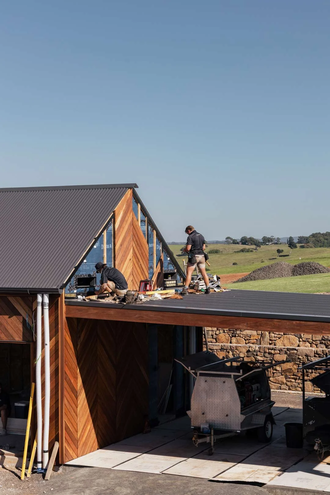 Construction workers installing or finishing wooden siding on a house with a metal roof in a rural area under a clear sky.