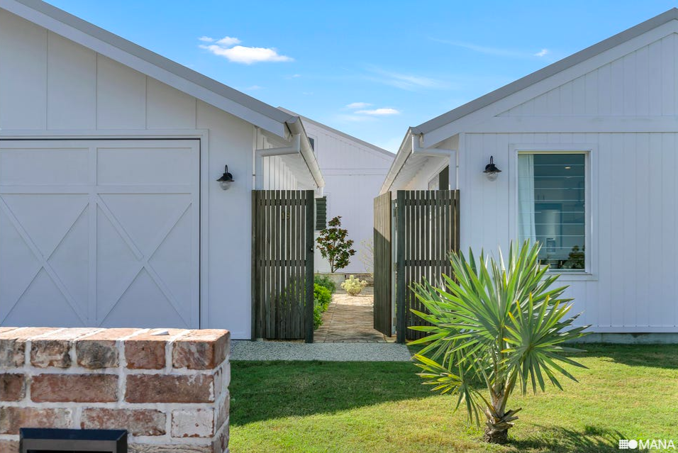 View of two white houses with gable roofs separated by a wooden gate, with a brick wall and green lawn in the foreground and a blue sky in the background.