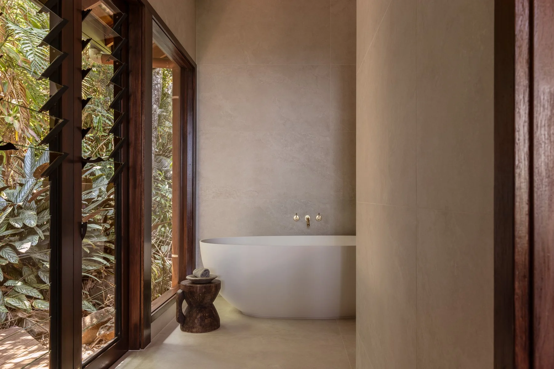 Modern bathroom with a free-standing bathtub next to a large window with wooden framing, overlooking lush green foliage outside. There is a small wooden stool with rolled towels beside the bathtub, and the walls are beige with minimalist fixtures.