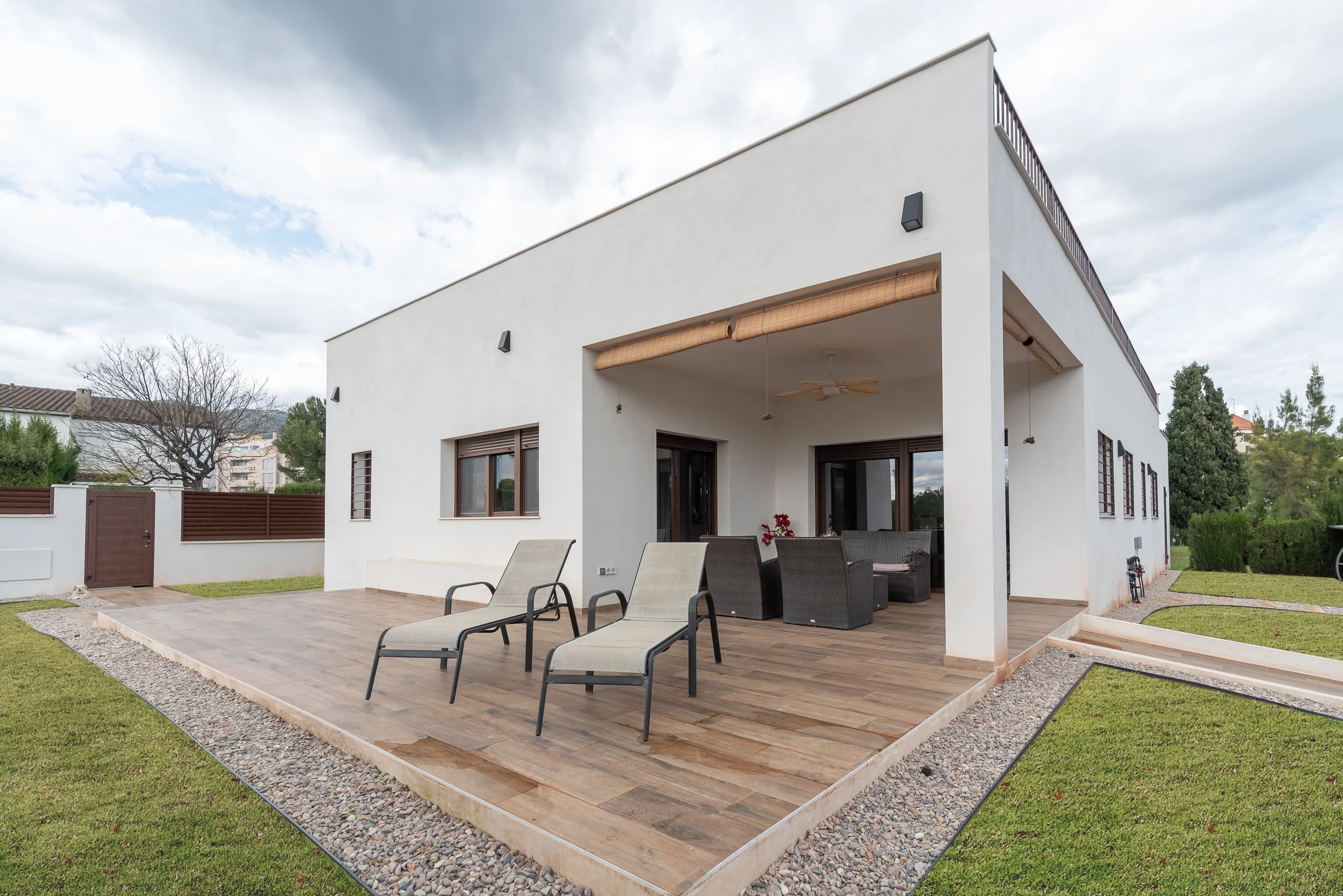 Proyecto de esvibar arquitectes. Casa moderna de dos pisos con terraza en el segundo nivel, área de descanso con sillones y toldo en la parte trasera, rodeada de césped y árboles, en un día soleado.