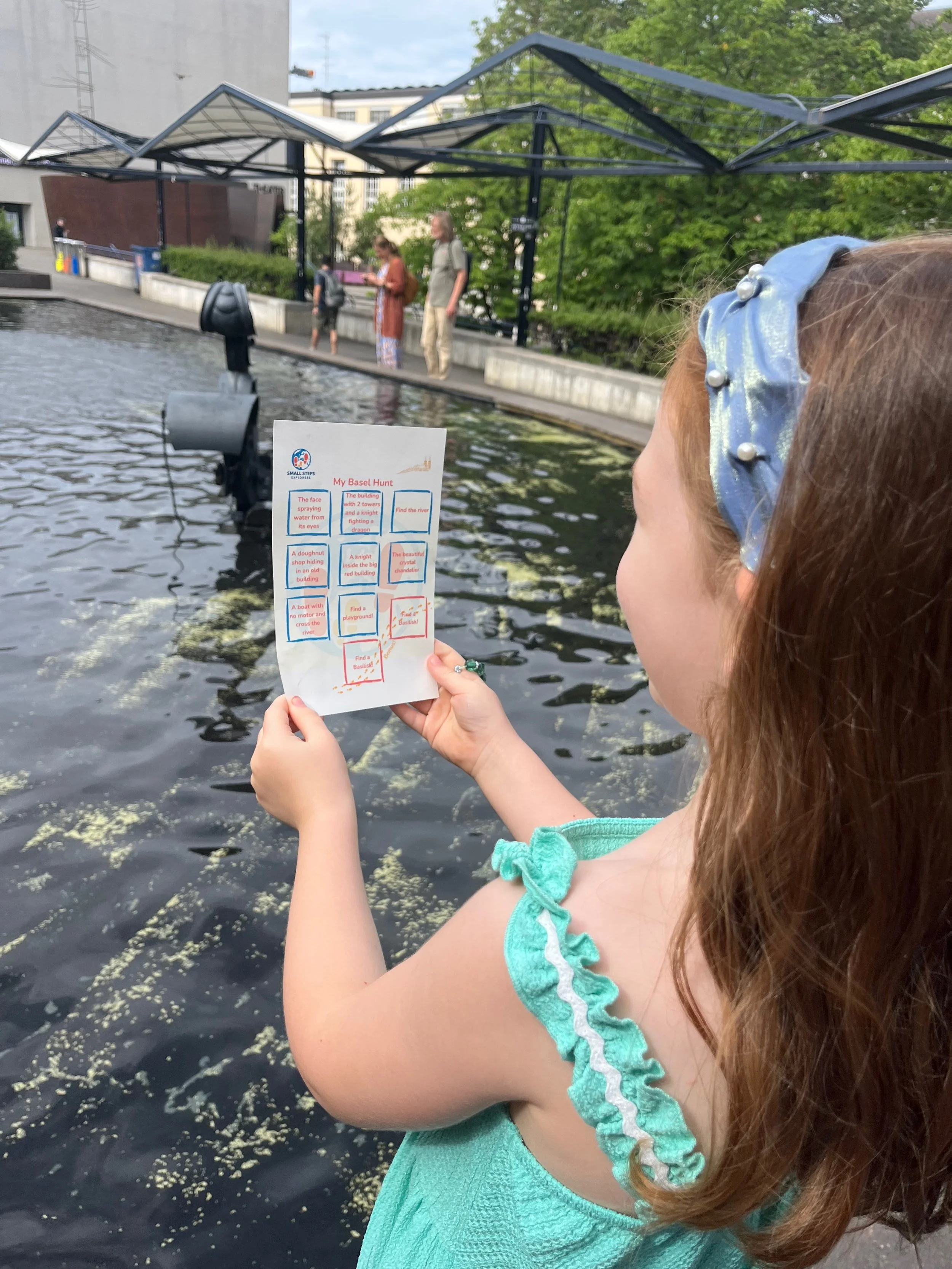 Child reading a Basel scavenger hunt sheet beside a city fountain — Small Steps Explorers