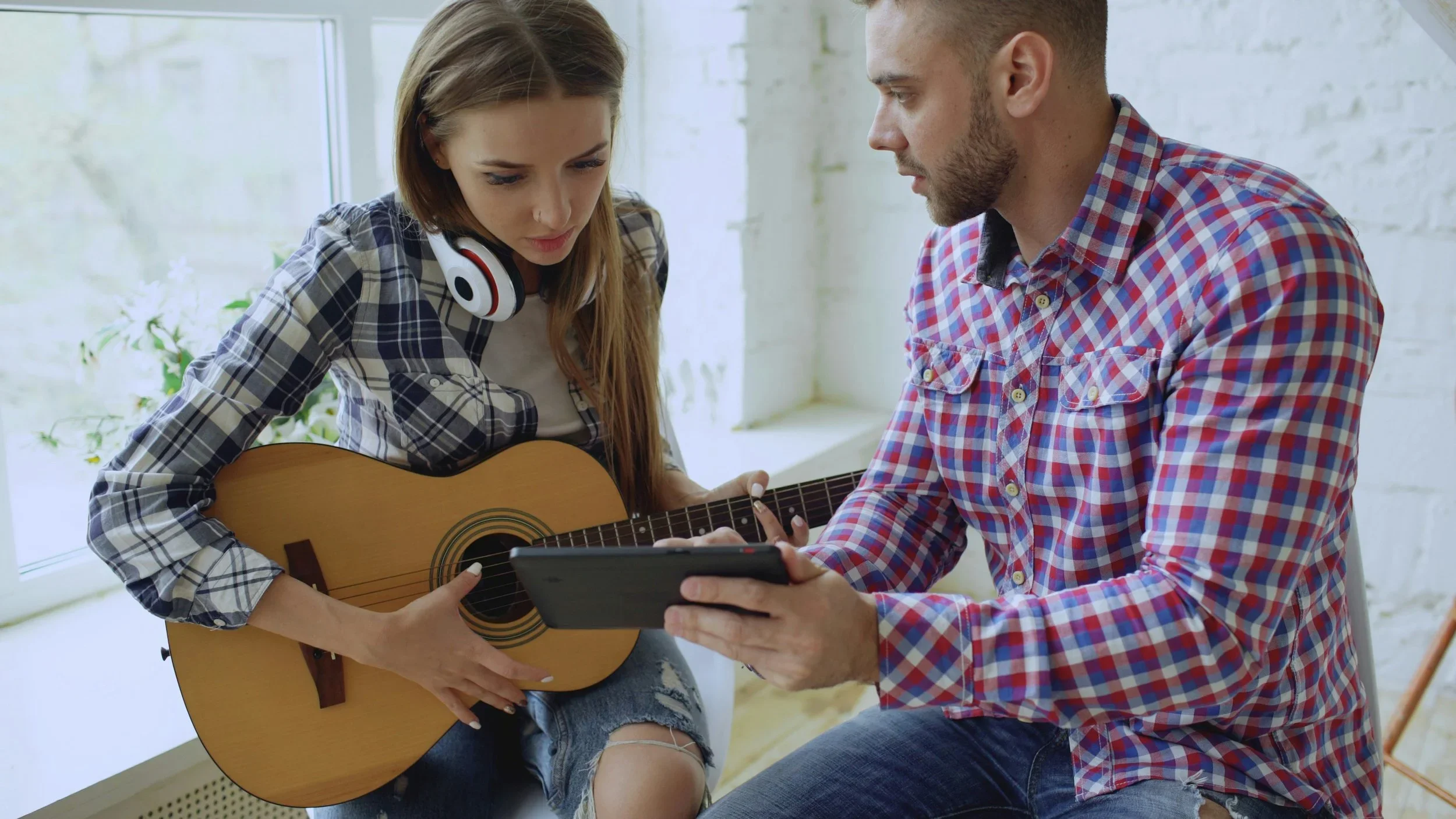 Une jeune femme avec des écouteurs autour du cou joue de la guitare pendant qu'un homme lui montre quelque chose sur une tablette, dans une pièce lumineuse avec une fenêtre en arrière-plan.