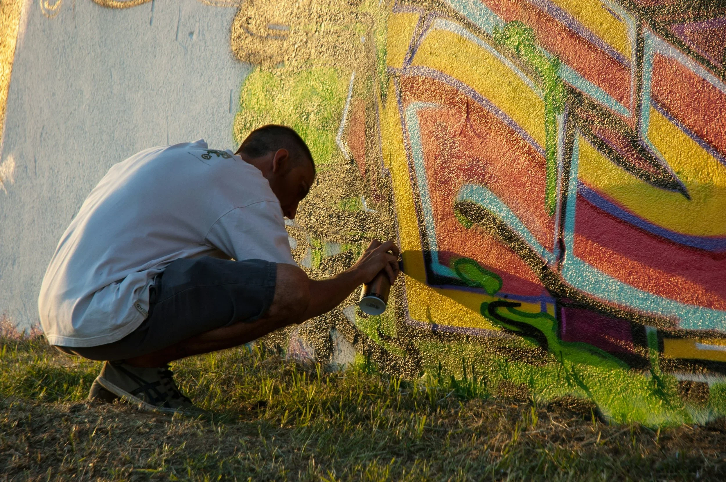 Un homme dessine un graffiti coloré sur un mur, avec de la peinture en spray, en position accroupie, au coucher du soleil.