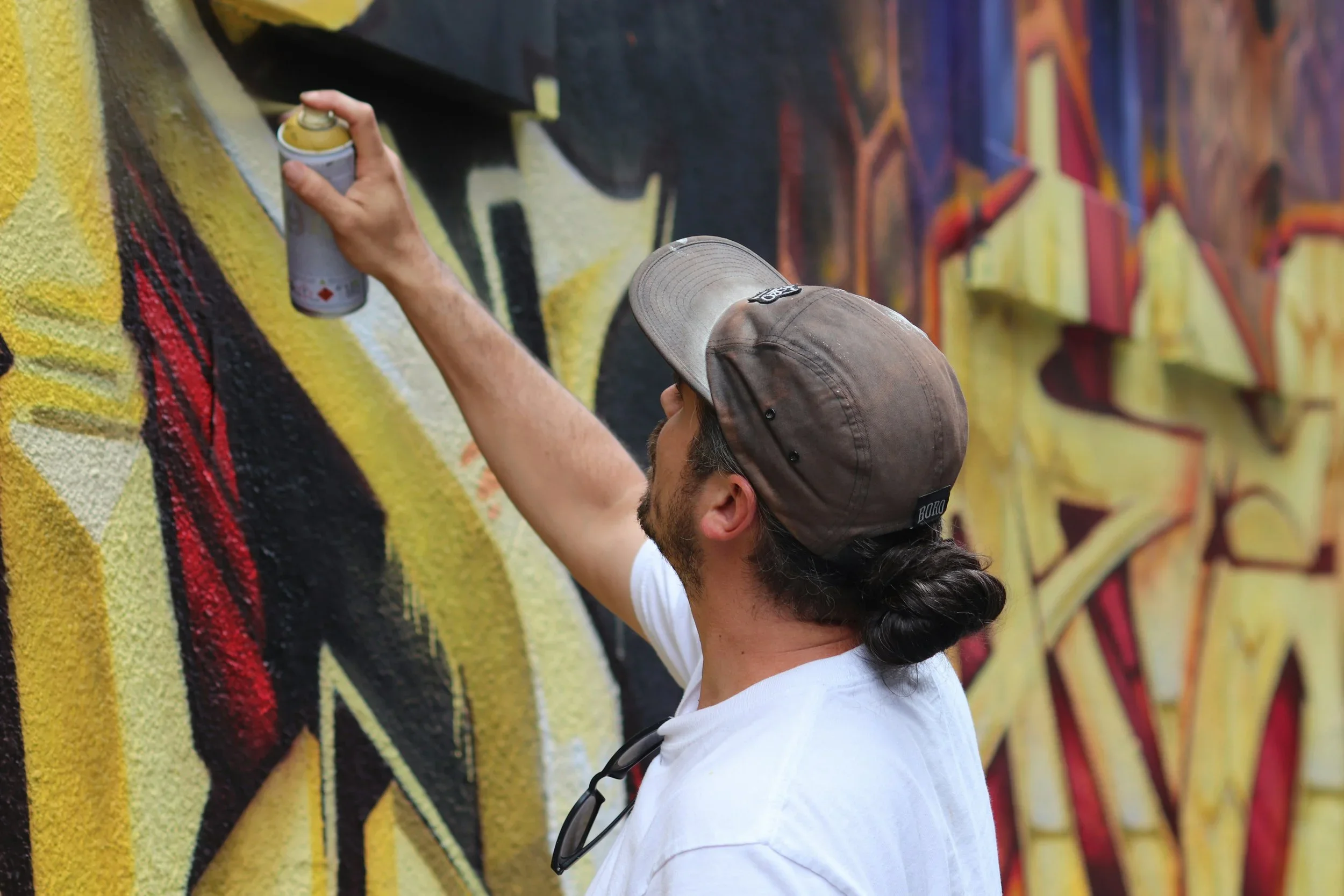 Un homme portant une casquette grise, une chemise blanche et des lunettes accrochées à son cou, utilise une bombe de peinture en spray pour créér ou colorier un graffiti coloré sur un mur.
