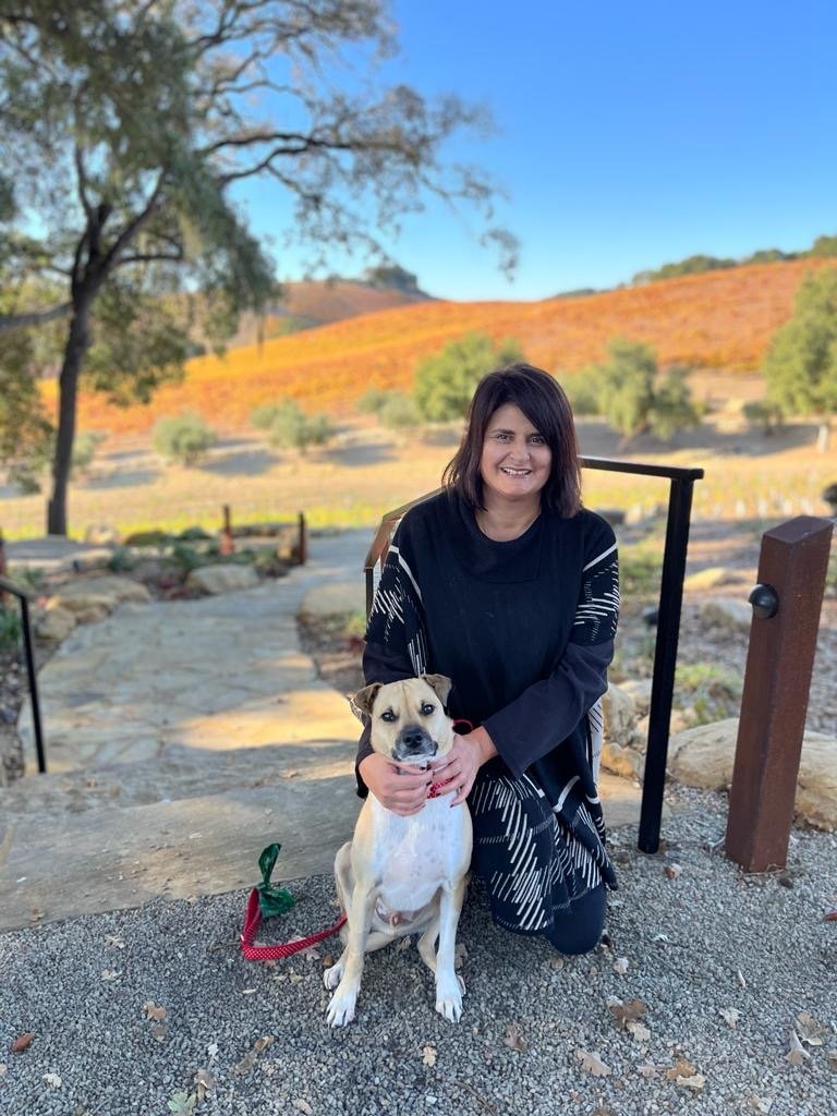 A woman kneeling next to a tan and white dog outdoors on a stone pathway during fall, with trees and hills with orange and green foliage in the background.