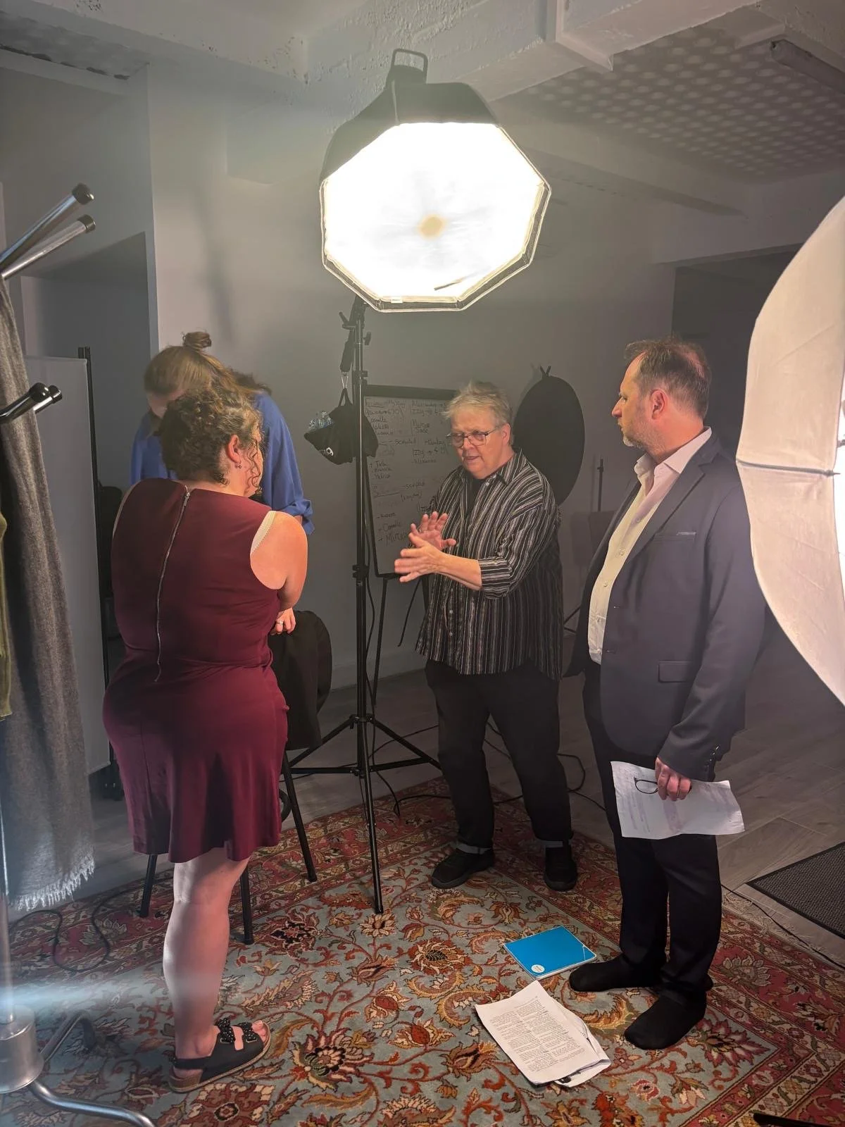 A group of people in a photography studio engaged in a discussion, with professional lighting equipment, a whiteboard, and papers on the floor.