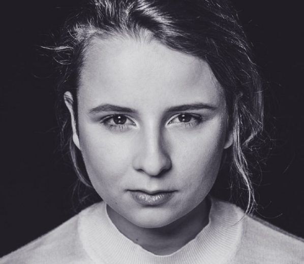 Close-up black and white portrait of a young woman with her hair pulled back, wearing a light-colored top, looking directly at the camera with a serious expression.