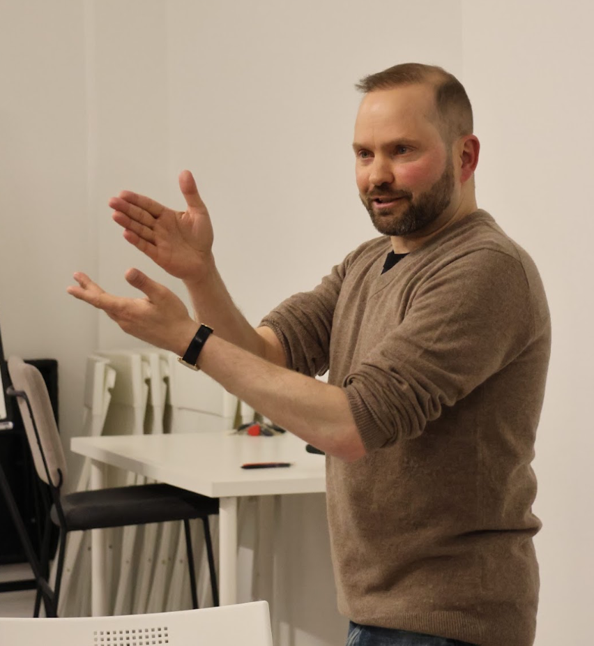 A man with a beard and short hair gestures with his hands while speaking in a room with white walls and white chairs.