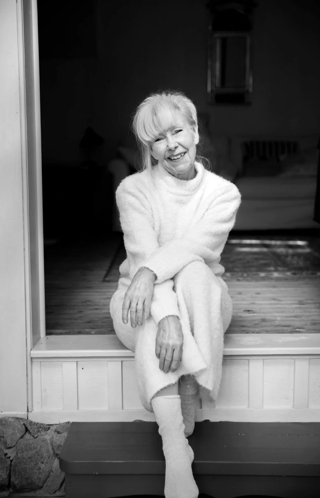An elderly woman sitting on a step inside her home, smiling at the camera. She is wearing a cozy, fluffy sweater and long socks, with her hair tied back.