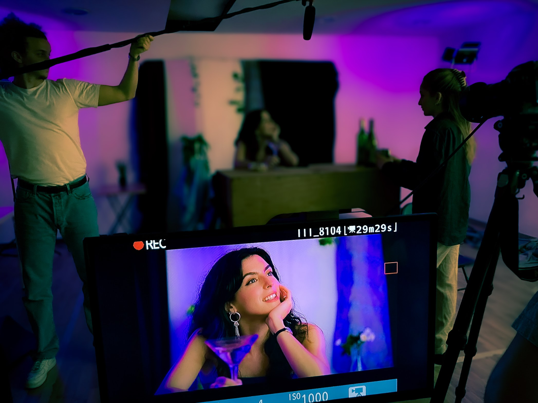 A behind-the-scenes shot of a video or photo shoot, showing a woman on a monitor in the foreground with a woman smiling and posing with a cocktail glass in the background.
