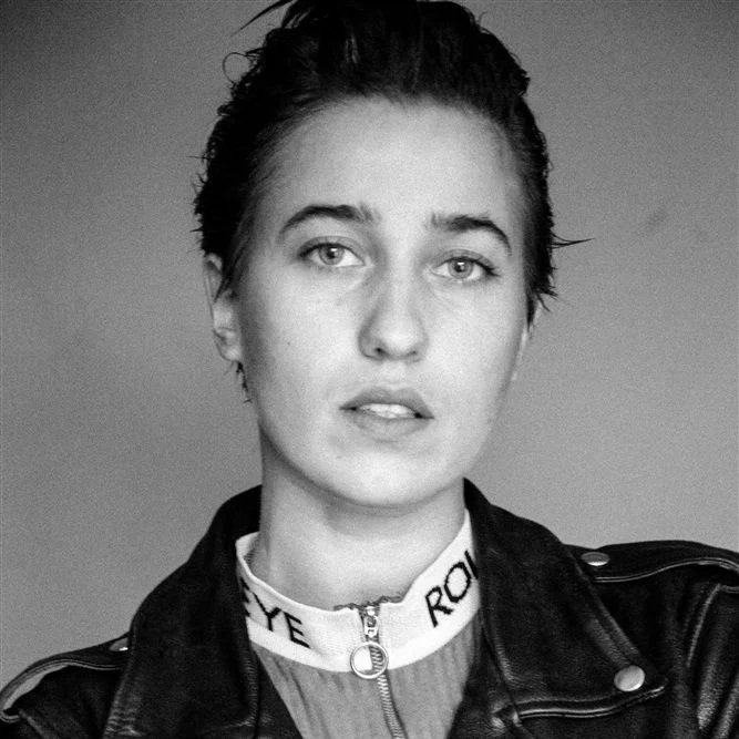 Black and white close-up photo of a young woman with short, wet hair wearing a leather jacket and a high-neck top.