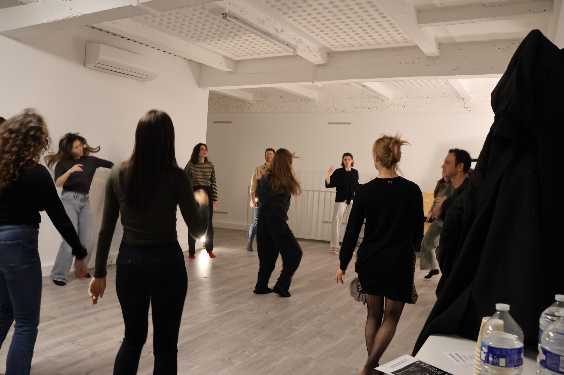 People participating in a dance or movement class in a spacious, well-lit room with white walls and wooden flooring.