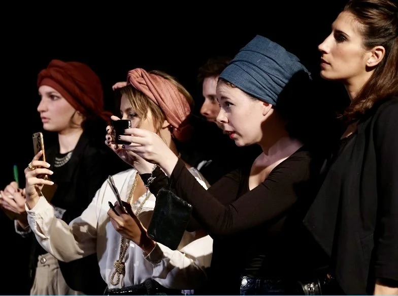 Group of women dressed fashionably, some taking photos or videos with smartphones, standing against a dark background.