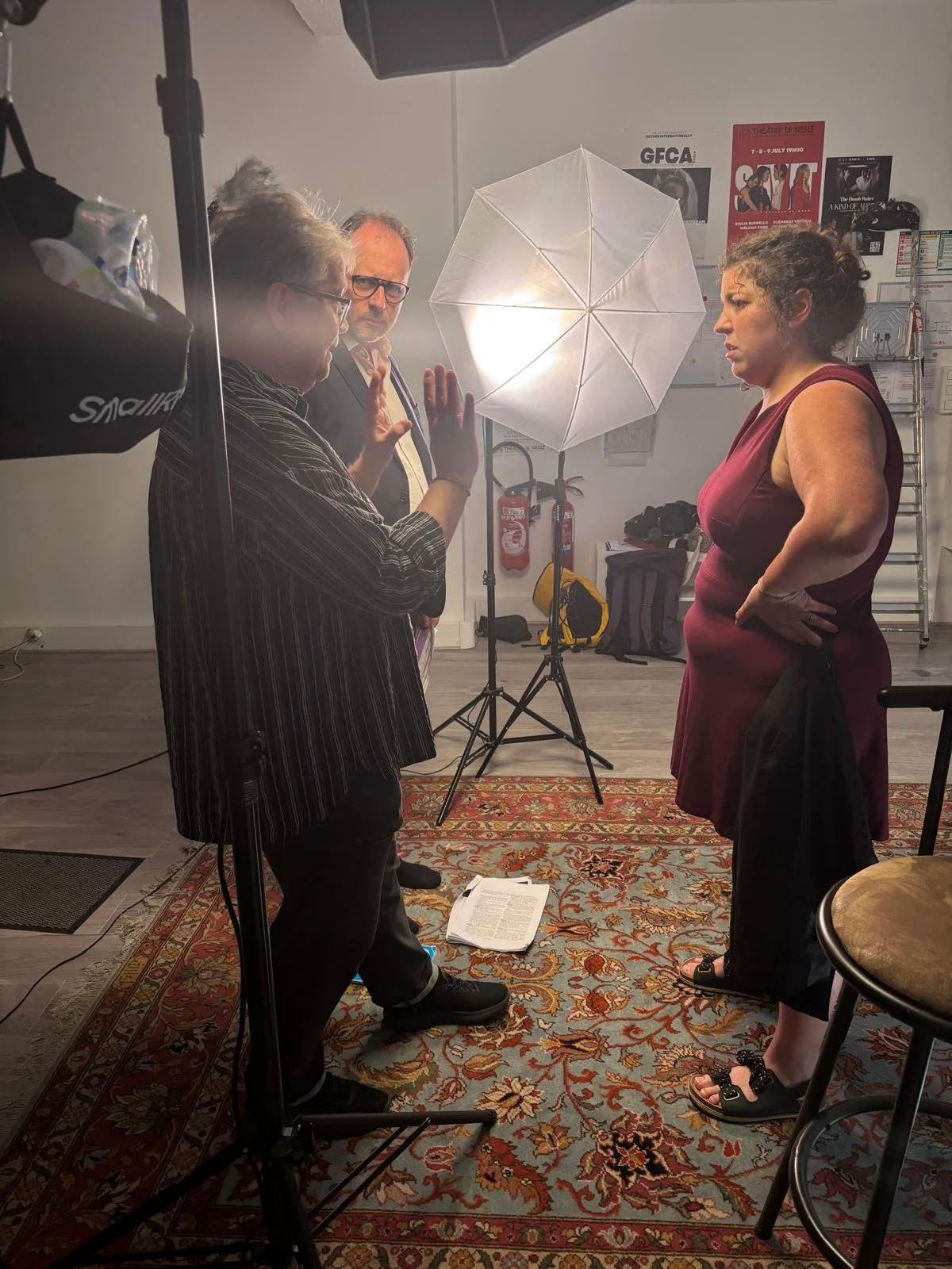 Three people in a photo shoot, with one woman standing and two men talking, surrounded by studio equipment including a light, an umbrella, and a rug on the floor.