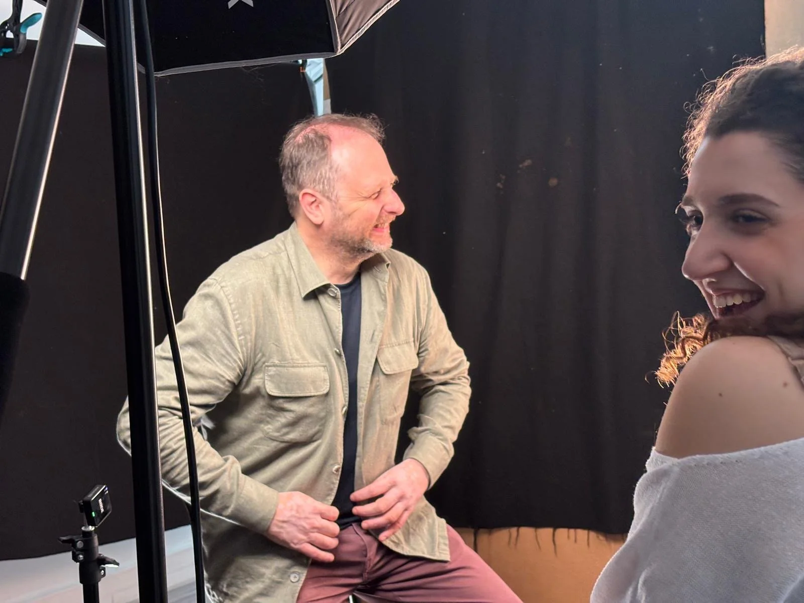 A man and a woman are seated indoors, smiling and laughing during a photoshoot or interview, with photography equipment visible.