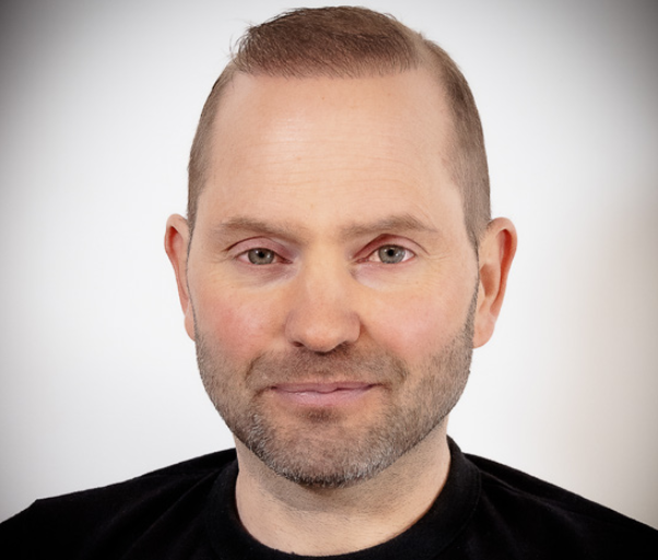 Close-up of a man with short brown hair and facial hair, wearing a black shirt, smiling slightly against a plain background.