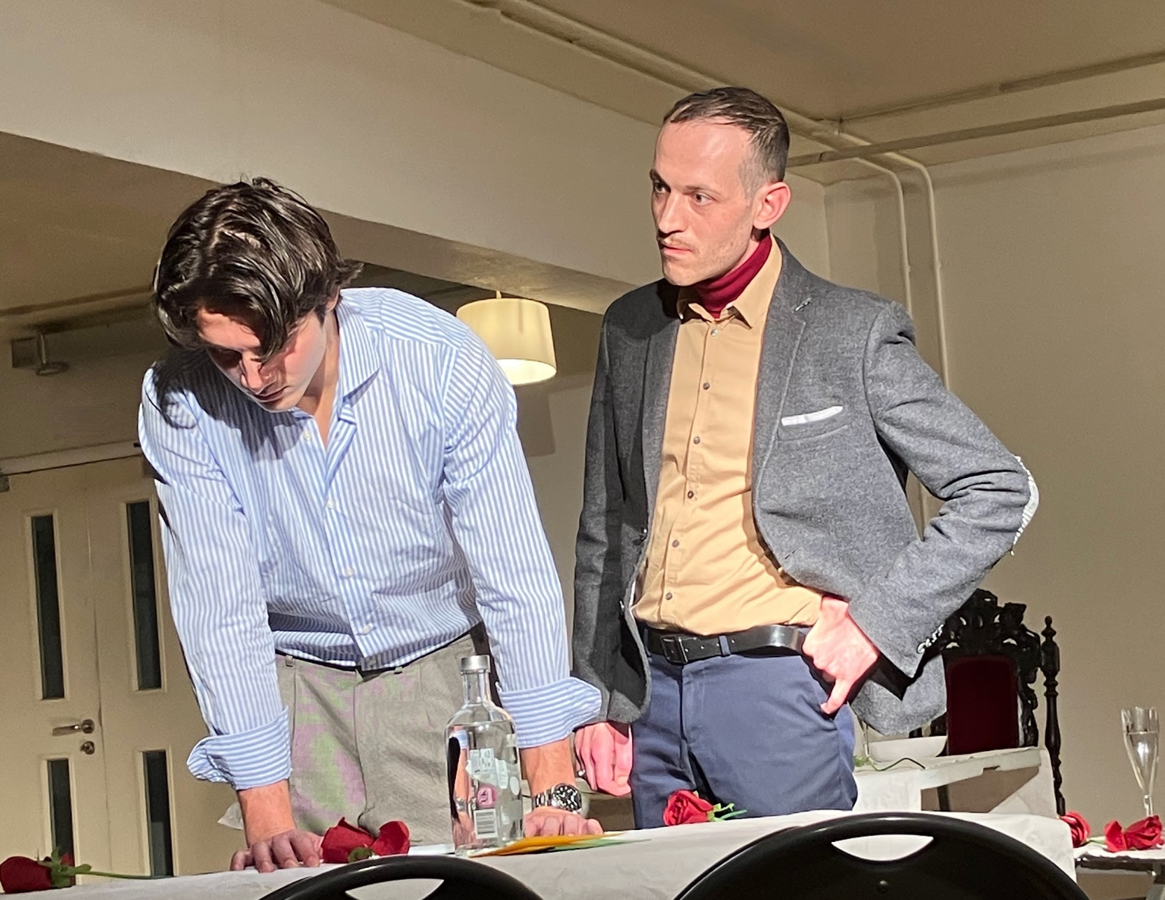 Two men standing at a dining table, one is leaning forward and the other has his hand in his pocket, with red roses on the table, a bottle of water, and glasses; indoor setting with beige walls and lighting.