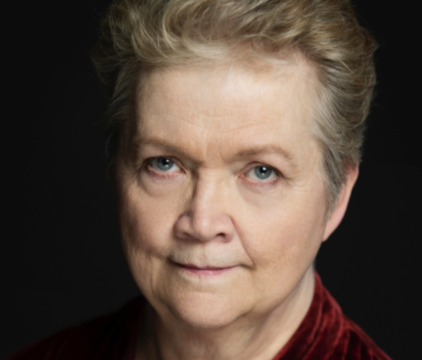Close-up portrait of an older woman with short blonde hair, blue eyes, and a slight smile, wearing a red top against a dark background.