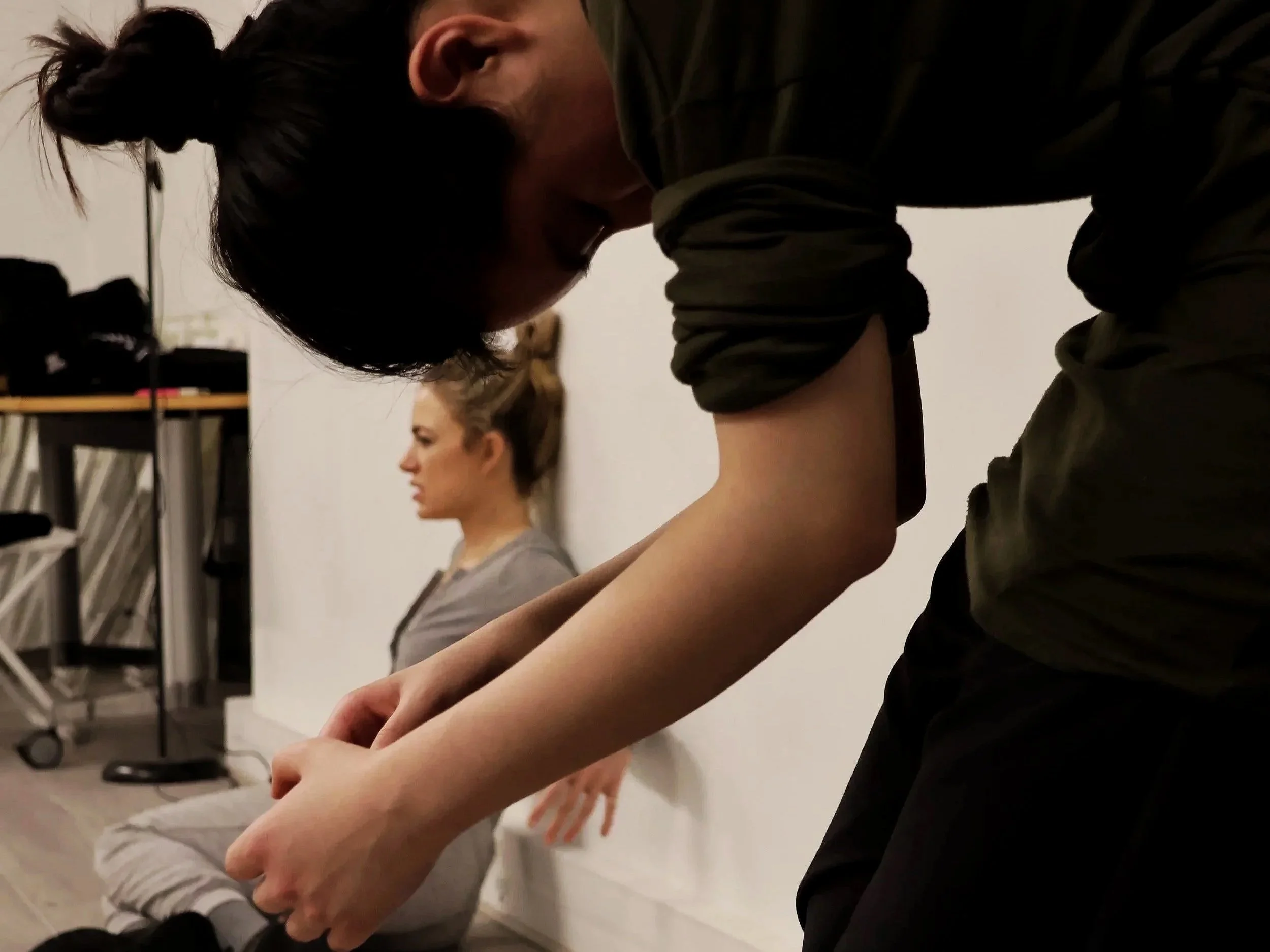 Two women kneeling with their heads bowed, engaging in a prayer or meditation, in a room with shelves and clothing in the background.