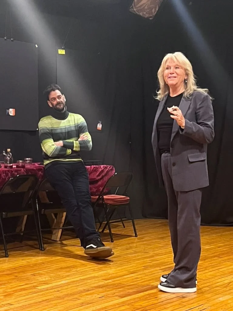 A woman in a gray suit is speaking or presenting, while a man in a green and black striped sweater stands nearby with crossed arms, listening in a room with black walls and wooden floors.