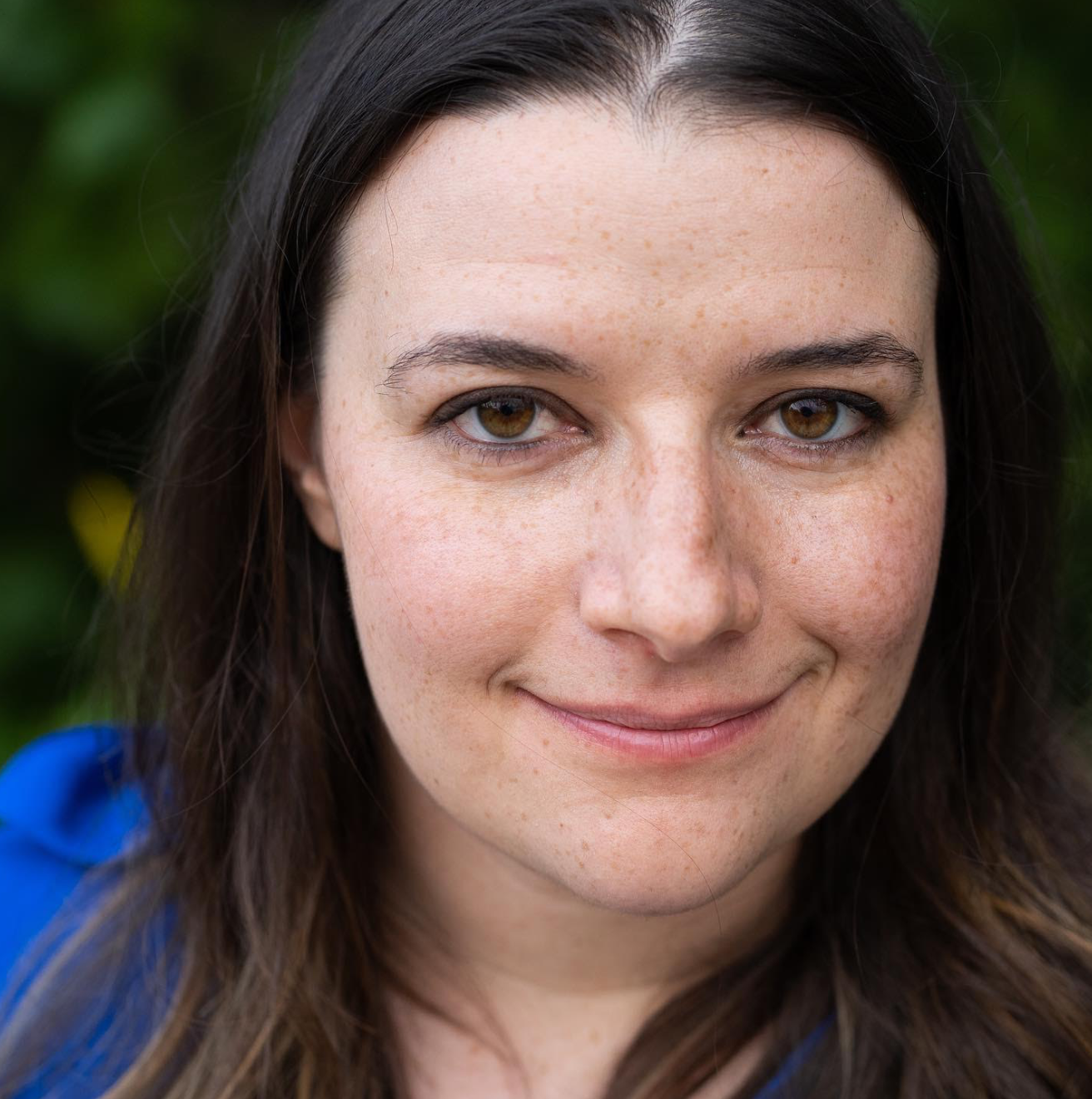 Close-up of a woman with long dark brown hair, fair skin, freckles, and brown eyes, smiling slightly, wearing a blue top, outdoors with blurred green foliage in the background.