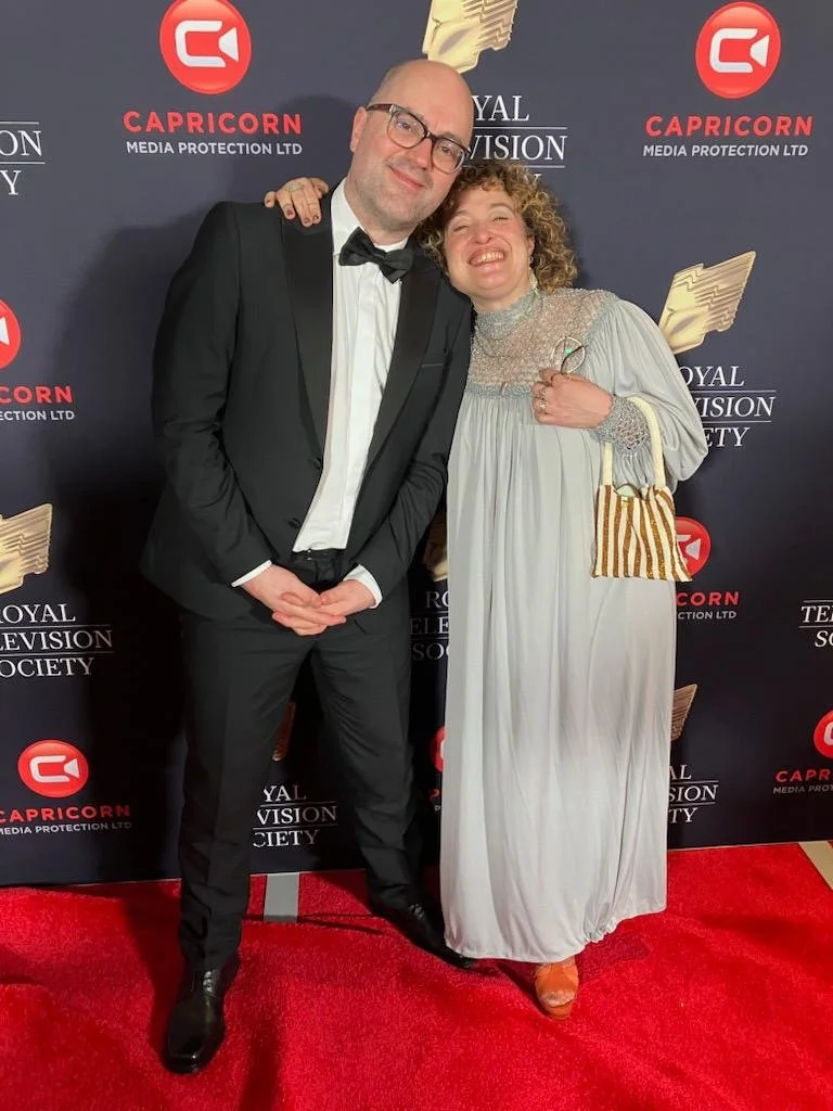 A man in a black tuxedo and glasses and a woman in a long white dress are smiling and standing close together on a red carpet at an event.
