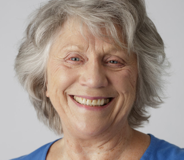 Close-up of a smiling elderly woman with gray hair and light skin, wearing a blue top.