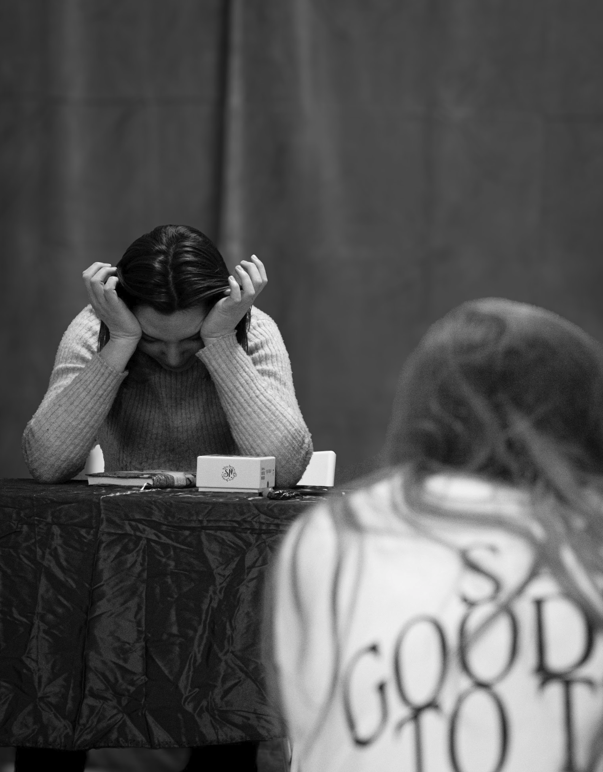A woman sitting at a table with her head in her hands, appearing distressed or upset. There is a box and a candy bar on the table in front of her, and another person is partially visible in the foreground. The setting seems to be indoors, with a plain wall in the background.