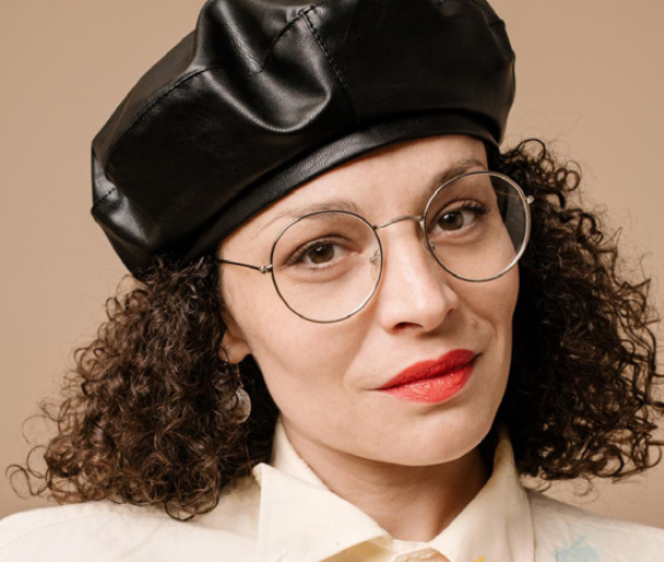 A woman with curly hair wearing round glasses, red lipstick, a white shirt, and a black leather beret against a neutral background.