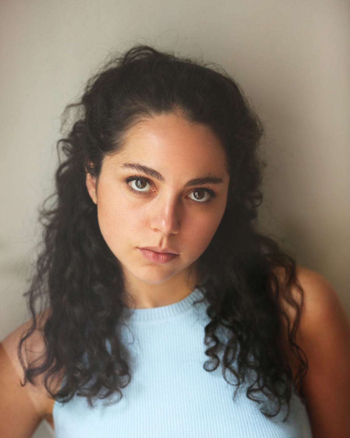 Close-up of a young woman with curly dark hair, blue eyes, wearing a light blue sleeveless top, looking directly at the camera against a plain light background.