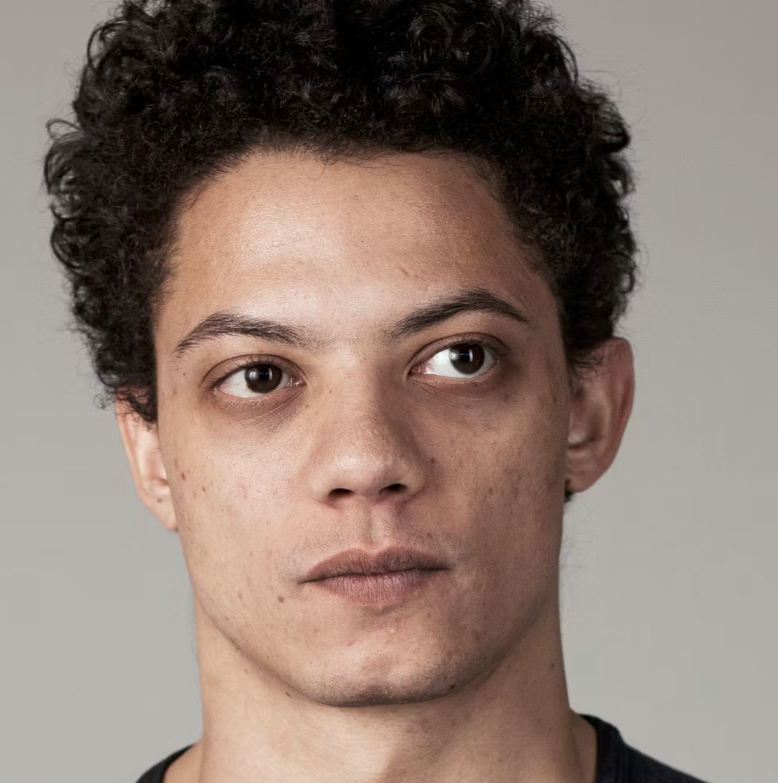 Close-up portrait of a young man with curly black hair and light facial skin, looking slightly to the right against a plain background.