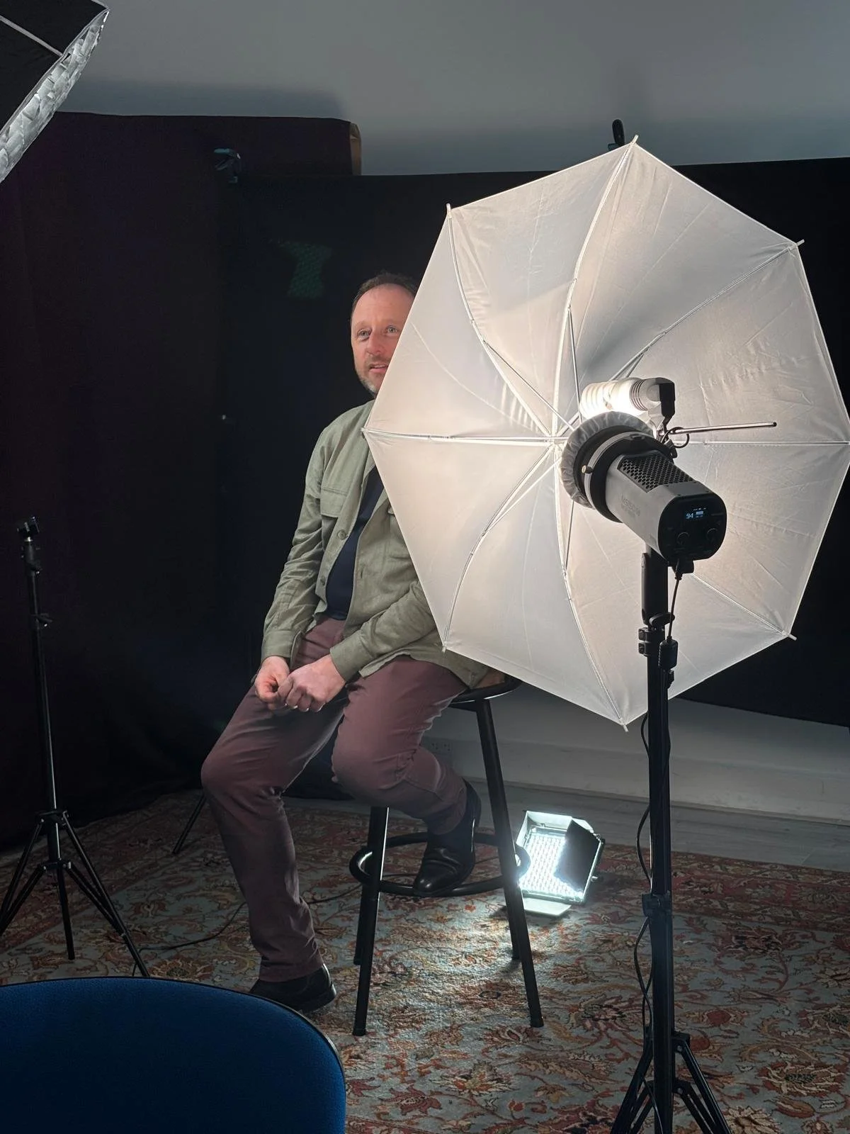 Man sitting on a stool in a photography studio with a white umbrella reflector and studio lighting equipment around him.