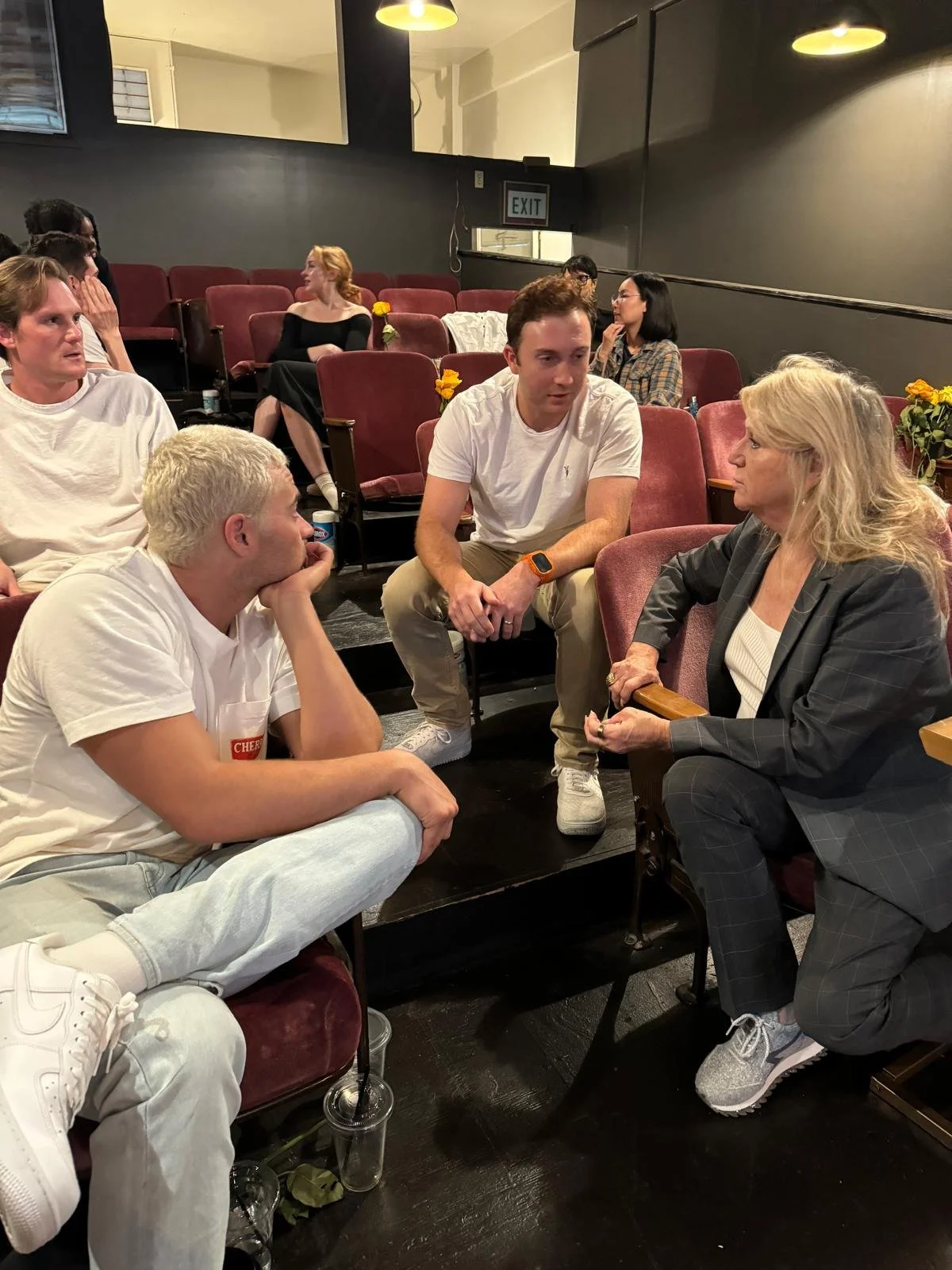 People engaging in conversation in a dimly lit theater or auditorium with red seats and dark walls.