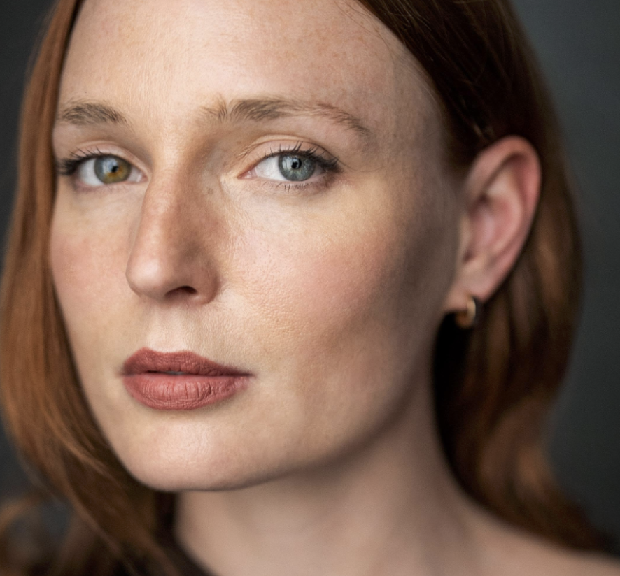 Close-up of a woman with red hair, blue and hazel eyes, and fair skin, looking at the camera with a neutral expression.