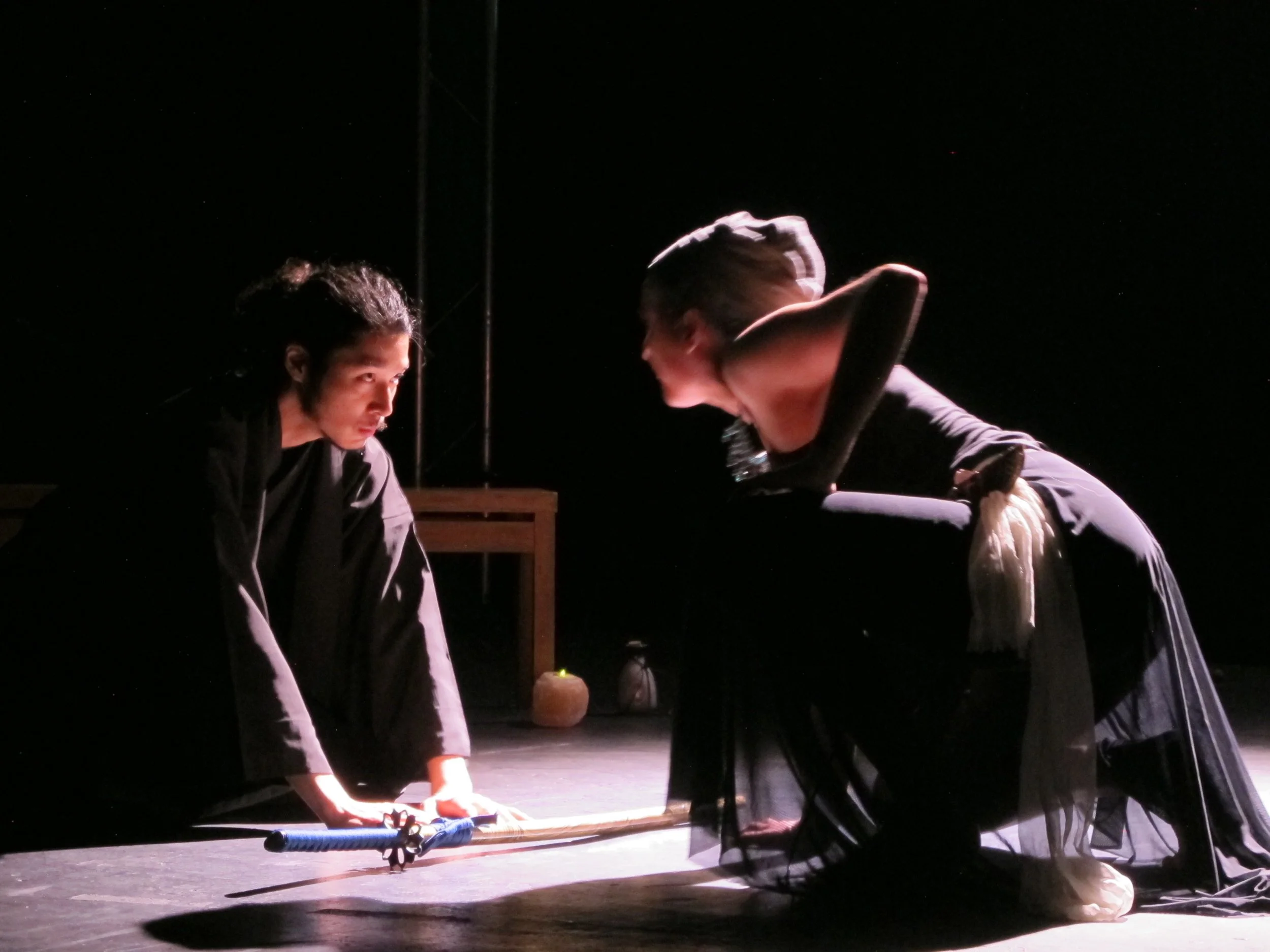 Two performers on stage in black costumes, leaning over a table with a prop, illuminated by spotlight, with dark background.