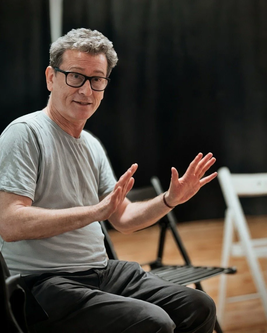 A man with gray hair, glasses, wearing a light gray t-shirt, is sitting on a black chair, gesturing with both hands, in a room with black walls and white chairs in the background.