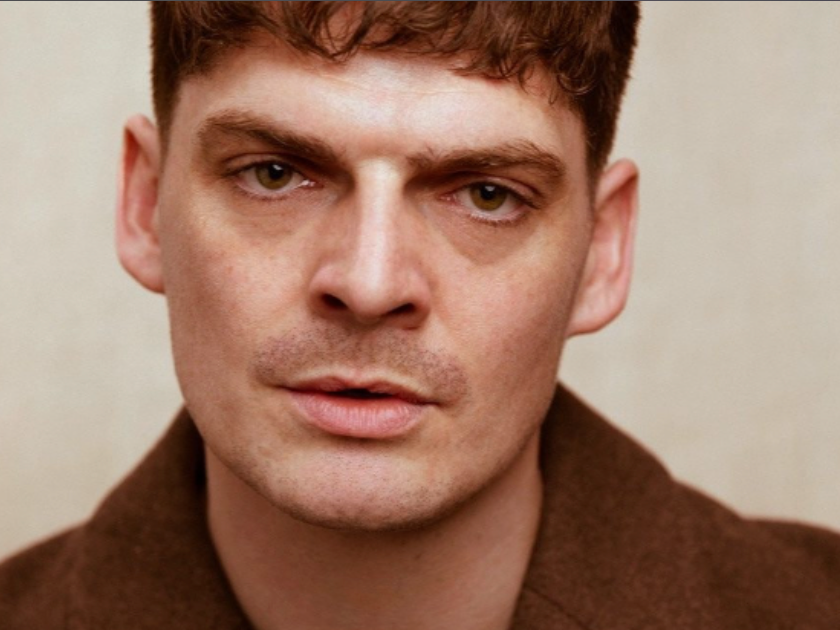Close-up of a young man with short reddish-brown hair, wearing a brown shirt, looking directly at the camera with a neutral expression.