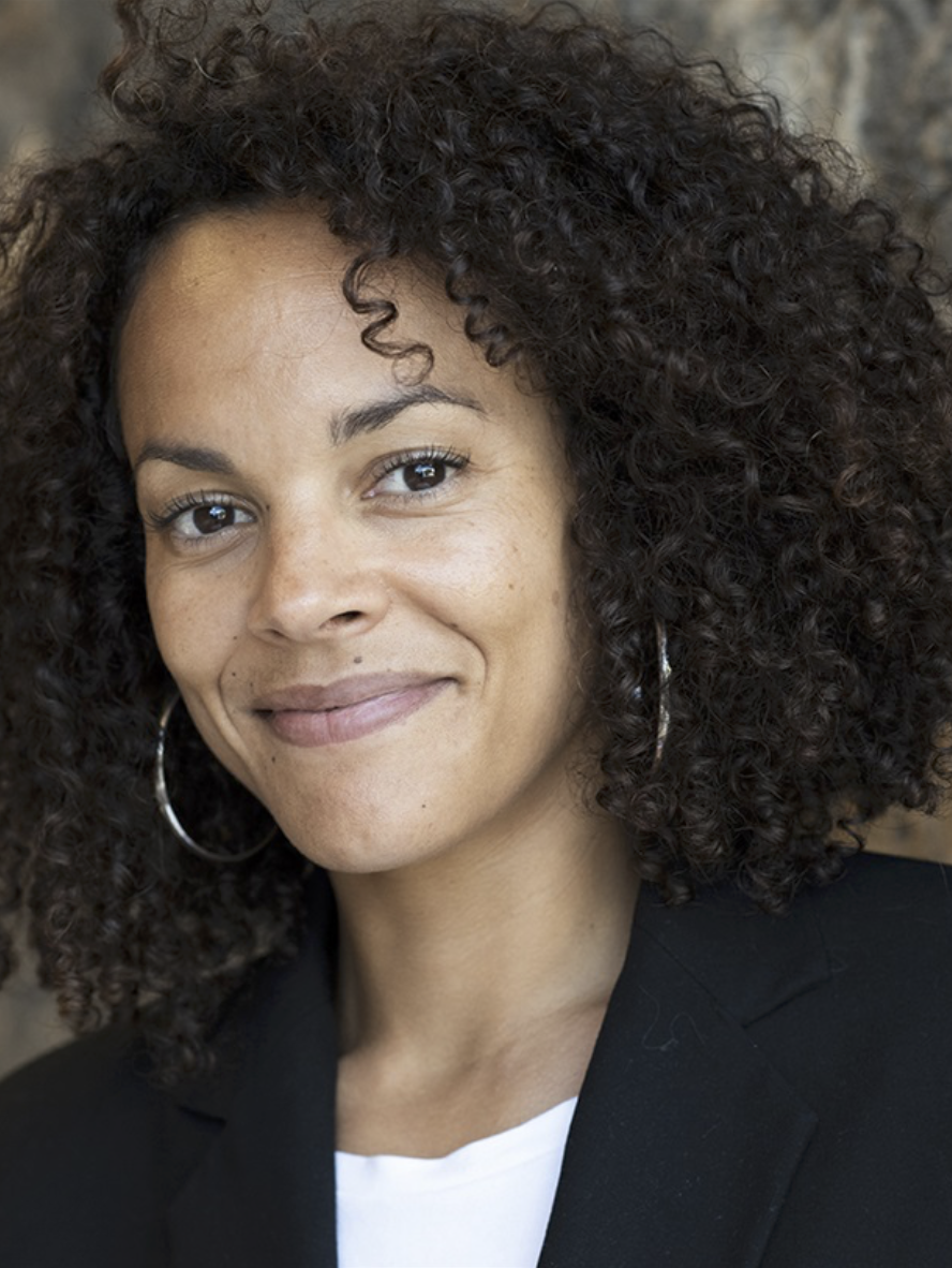 Close-up of a woman with curly dark hair, hoop earrings, wearing a black blazer and white top, smiling softly.