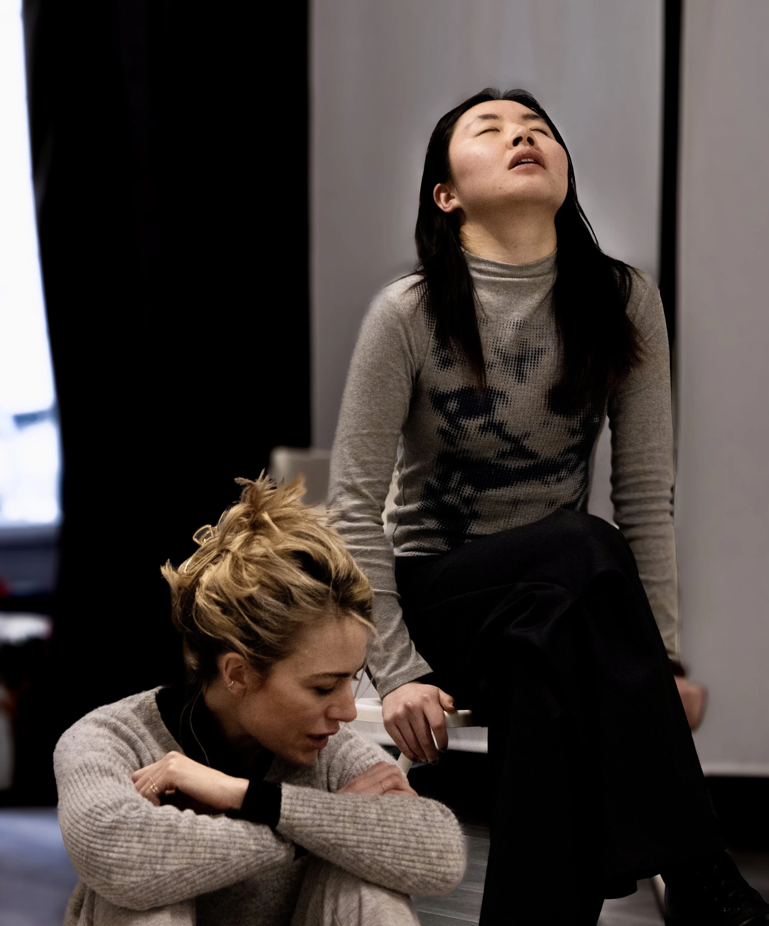 Two women in a room, one with platinum blonde hair in a bun and the other with long black hair, both engaged in individual activities.