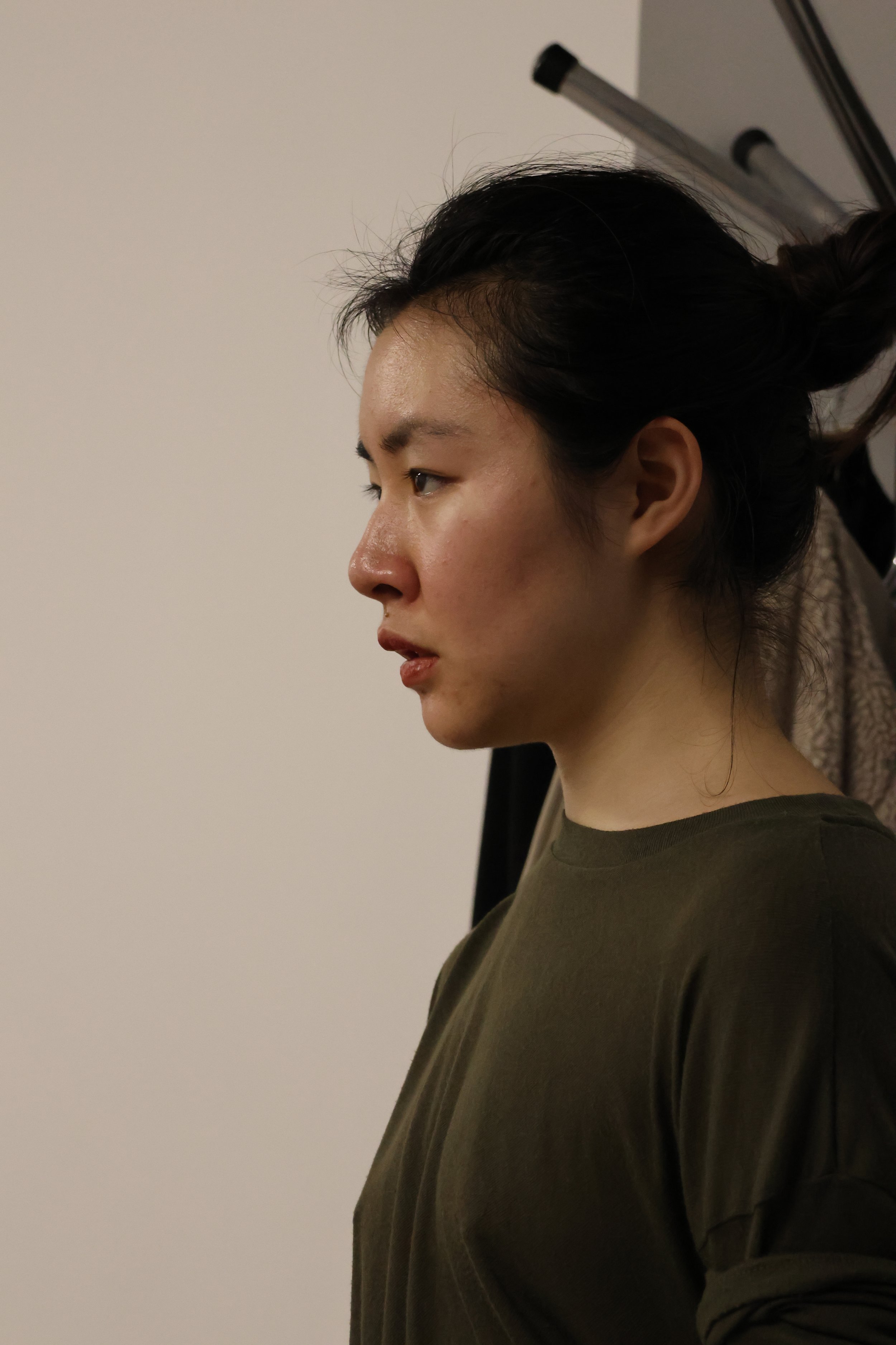 A woman with dark hair styled in a bun, wearing an olive-green shirt, seen in profile against a plain white wall.