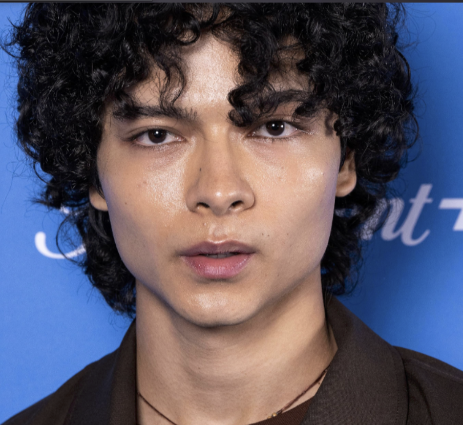 Close-up of a young man with curly black hair and light skin, looking directly at the camera, against a blue background.