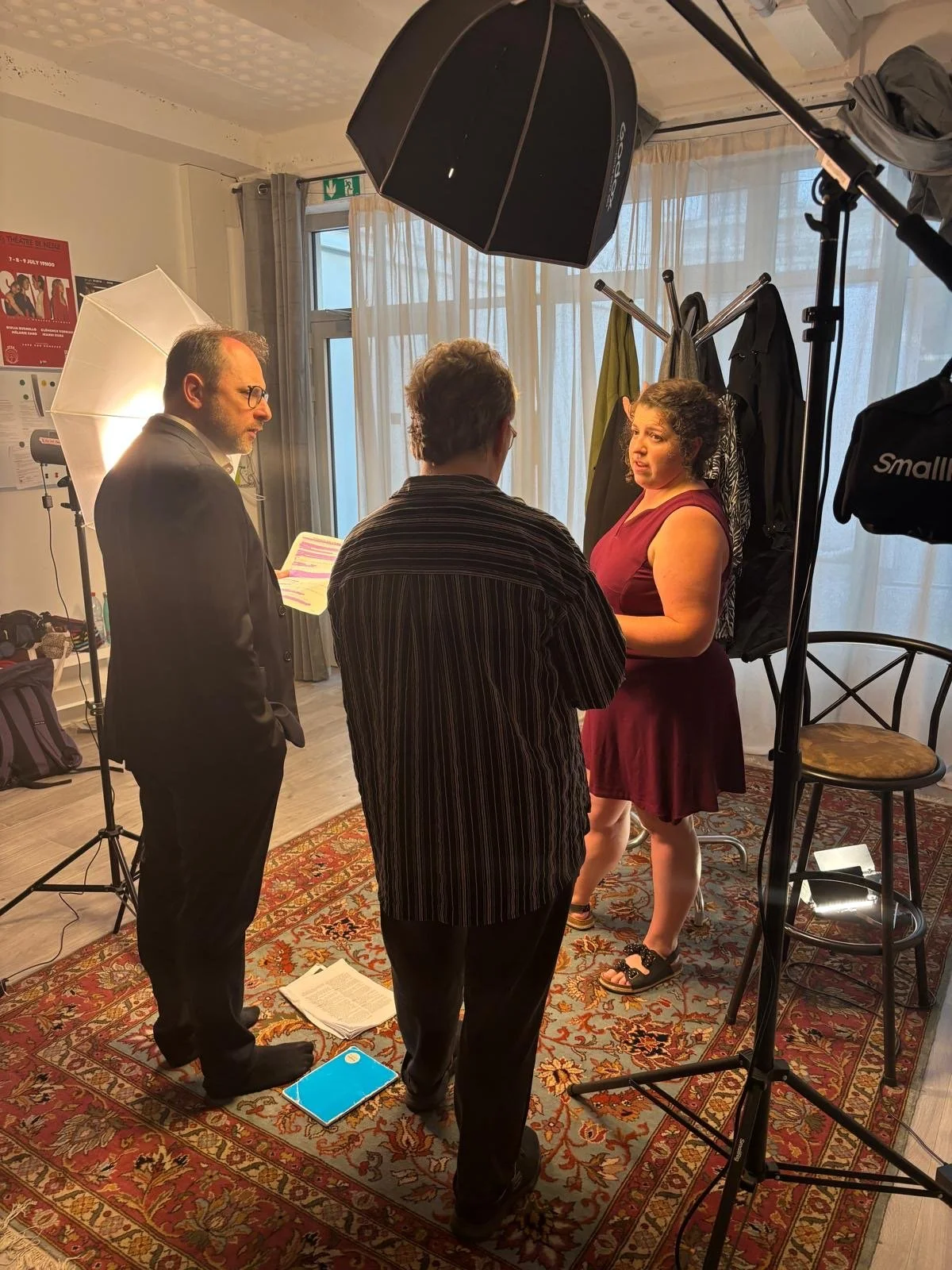 A woman in a burgundy dress participating in an interview or filming session in a room with curtains, surrounded by three men, one with glasses, holding papers, another with a striped shirt, and the third behind a camera setup with studio lights.