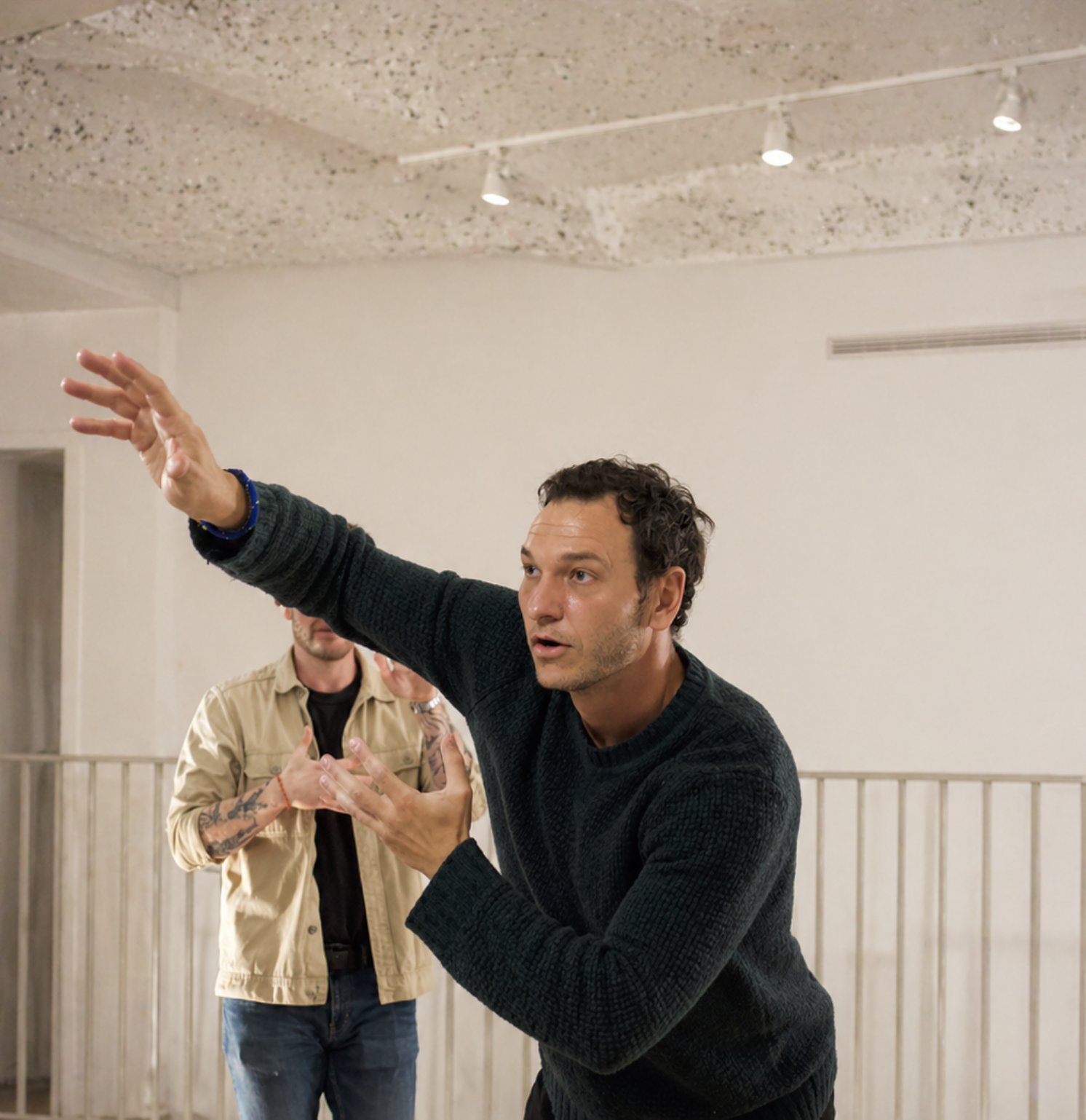 A man with dark curly hair wearing a black sweater gestures with his right arm extended in a room with a textured ceiling, two ceiling lights, and a white wall. Two other men are in the background, one wearing a beige shirt with tattoos on his arms and the other partially visible.