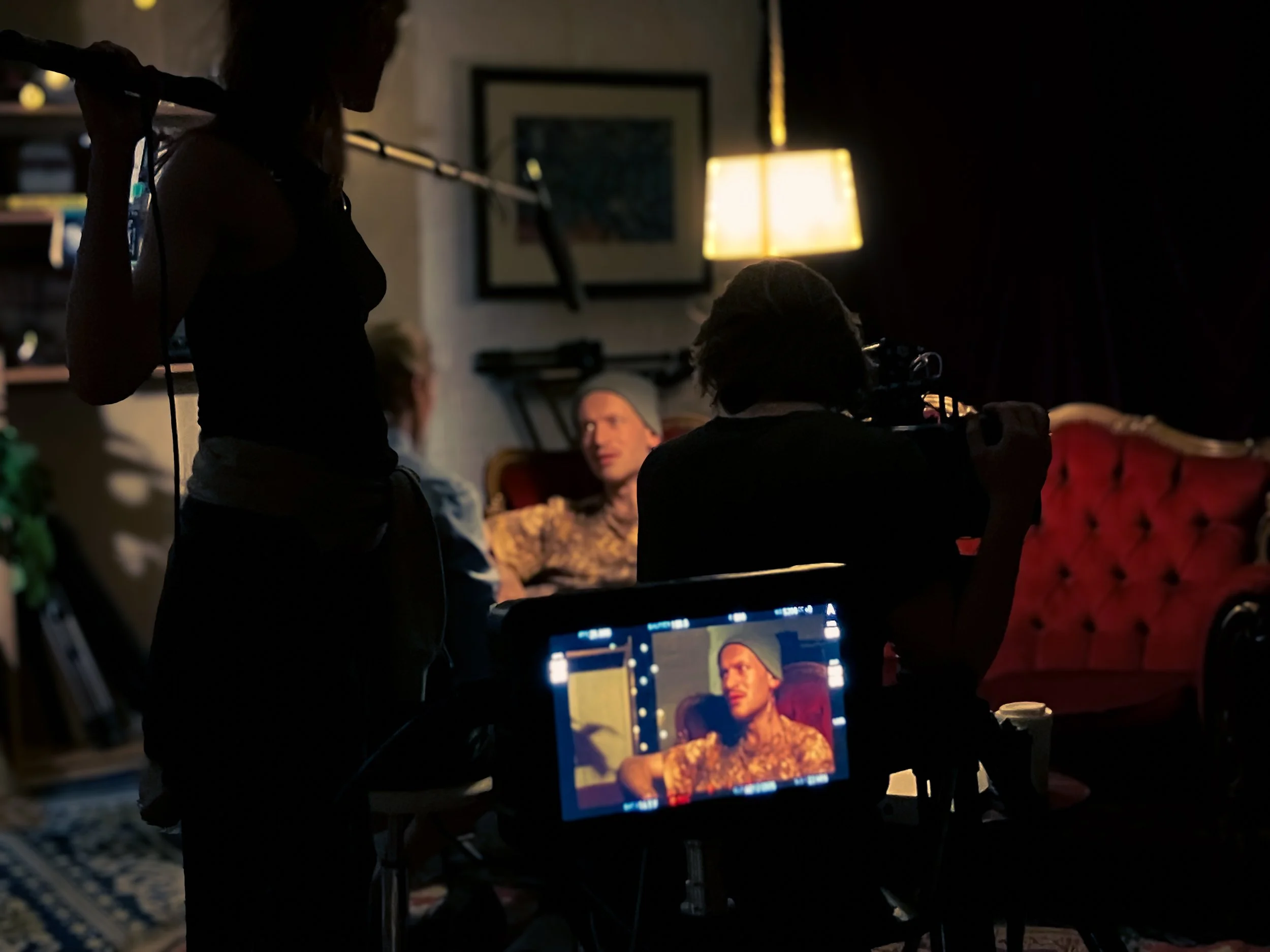Filmmaking scene in a dimly lit room with a director filming a woman sitting on a red velvet chair, a monitor displays her face while crew members work around. A table and a lamp are also visible.