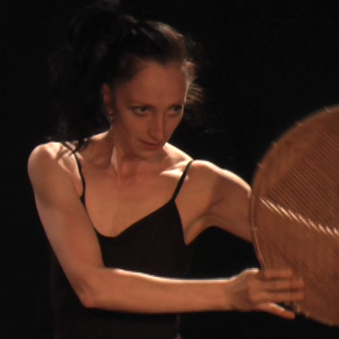 A woman with dark hair in a black spaghetti strap top holding a circular woven fan in front of a dark background.