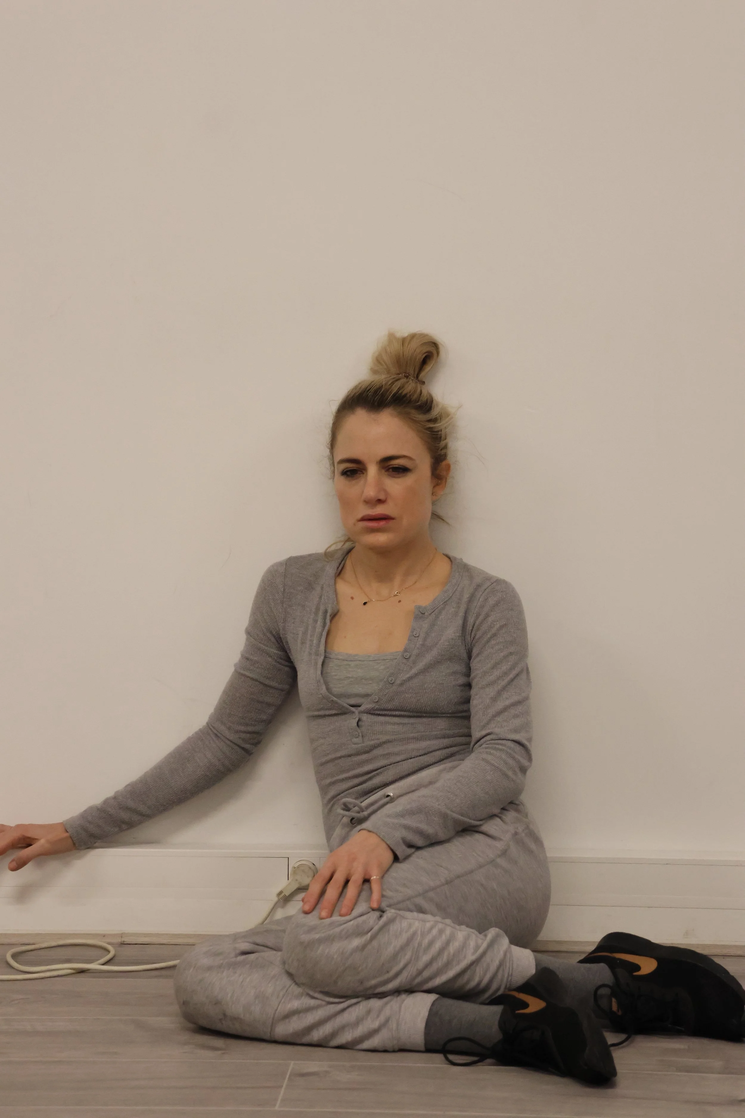 A woman with blonde hair in a top knot, wearing a gray cardigan and sweatpants, sitting on a light wood floor, leaning against a white wall with her eyes closed and a relaxed expression.