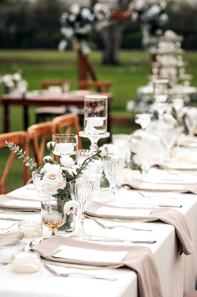 Une table de mariage élégante en plein air avec des verres, des papeteries, et un centre de table floral blanc, entourée de chaises en bois et un décor champêtre.