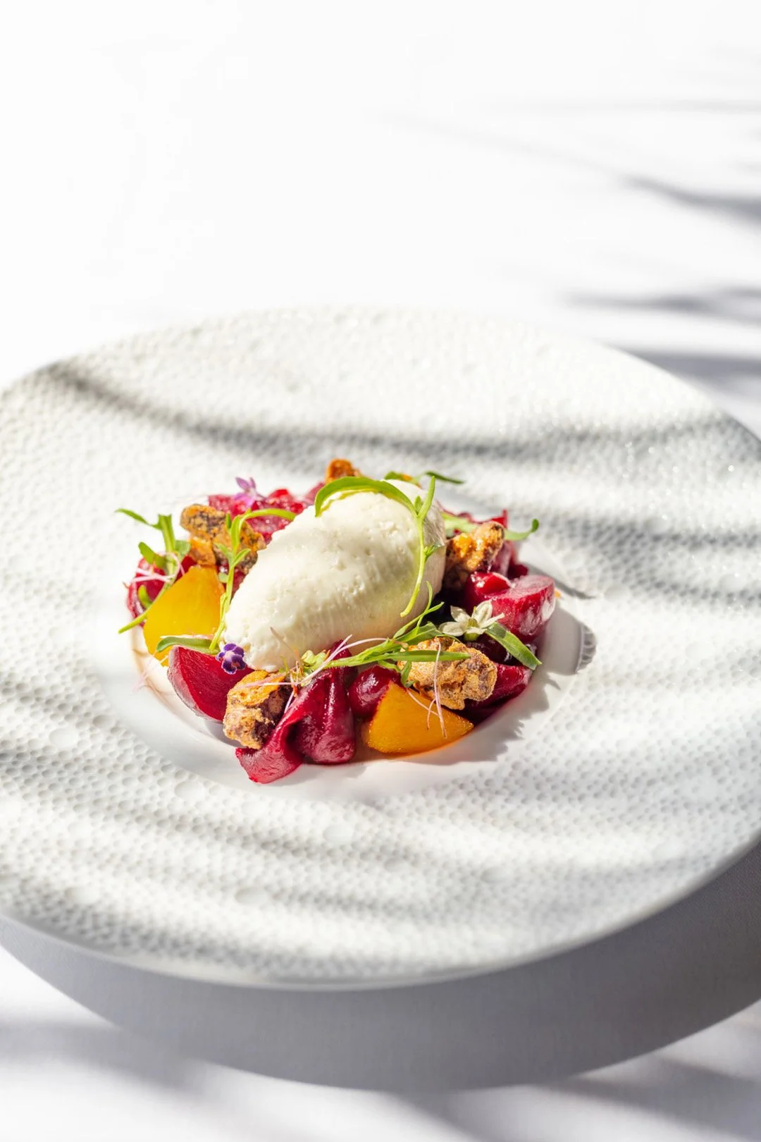 Un plat de dessert composé de morceau de fruits rouges, de pépites de chocolat, d'une boule de glace ou de sorbet, décoré de microgreens et de fleurs comestibles, présenté sur une assiette blanche avec un fond blanc