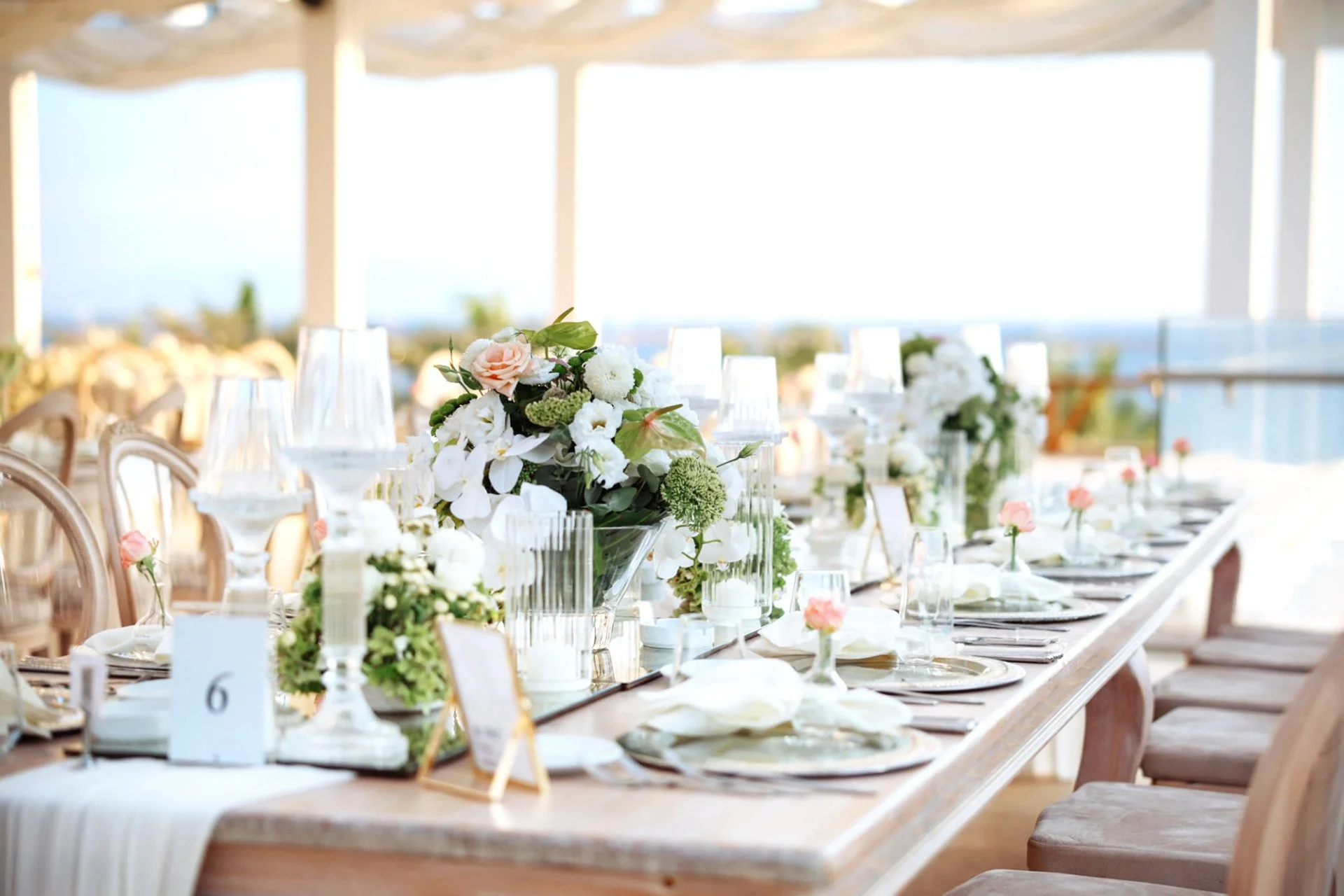 Une table de banquet élégante décorée de fleurs blanches et roses, avec des assiettes, des verres et des petits centres de table, dans un décor lumineux et aéré.